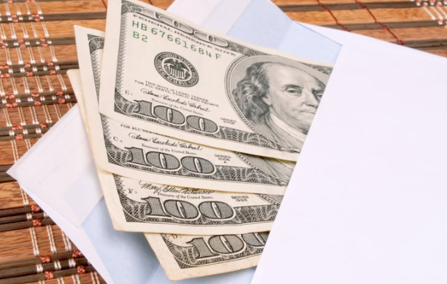 Three $100 bills partially visible in a white envelope, resting on a wooden surface.