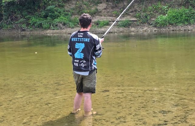 Person fishing in a shallow river, wearing a jacket and shorts.