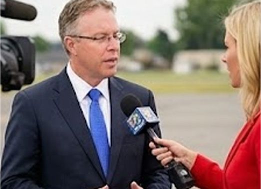 Man in suit being interviewed by a woman holding a microphone; outdoors.