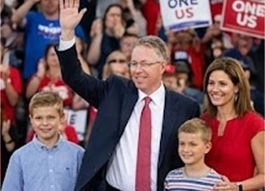 Man in suit waves to a crowd with family. Red and blue colors dominate.