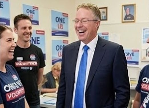 Man in suit laughing with a group of people, campaign posters in the background.