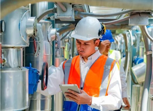 Man in hard hat and safety vest examines equipment with a tablet in an industrial setting.