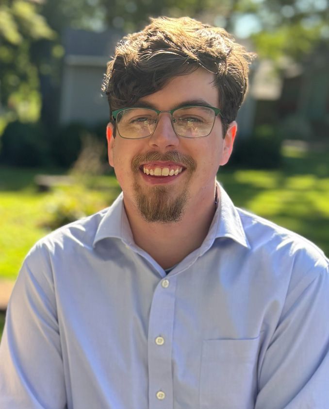 Man with glasses, light blue shirt, smiling outdoors.