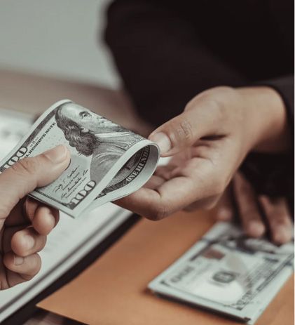 Hands exchanging a folded stack of $100 bills, another stack on a desk.
