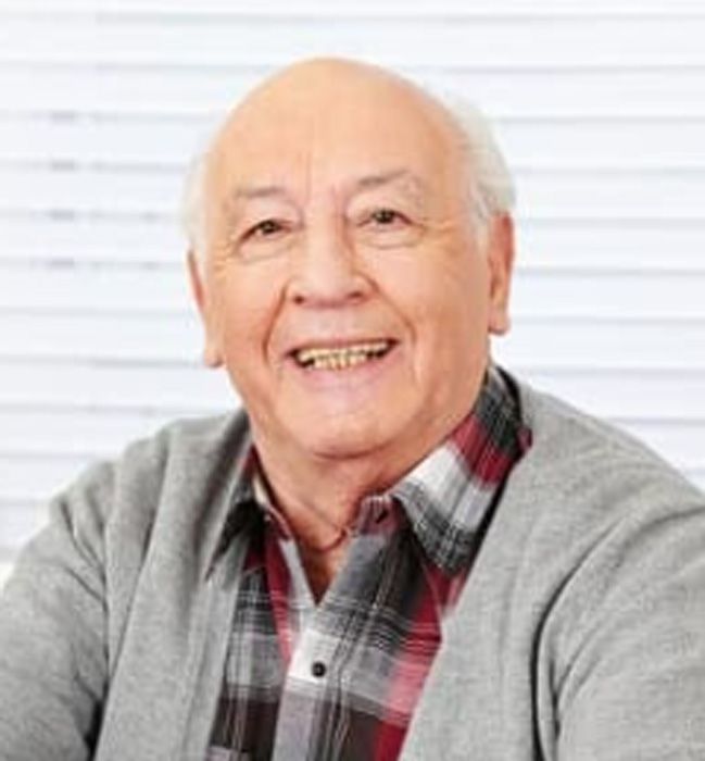 Smiling older person wearing a gray cardigan over a plaid shirt, in front of a window.