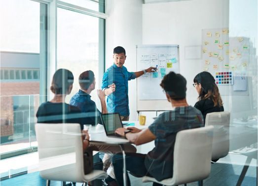 Group of people in a meeting. A man points at a whiteboard with sticky notes. Sunlight streams in from a window.