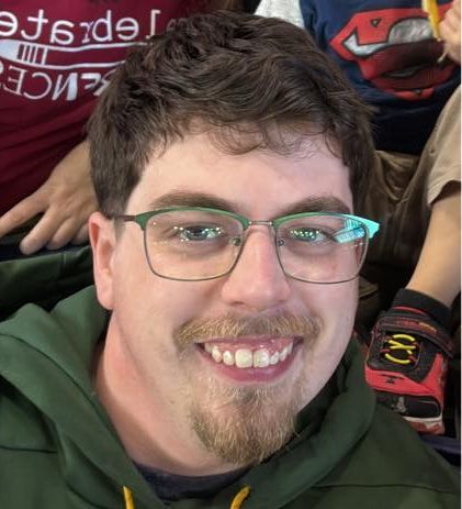 Man with green-framed glasses and a goatee smiles at the camera, wearing a green hoodie.