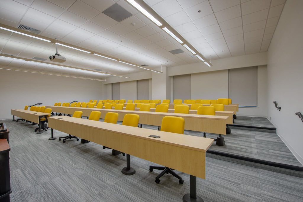 A large classroom with long tables and yellow chairs.