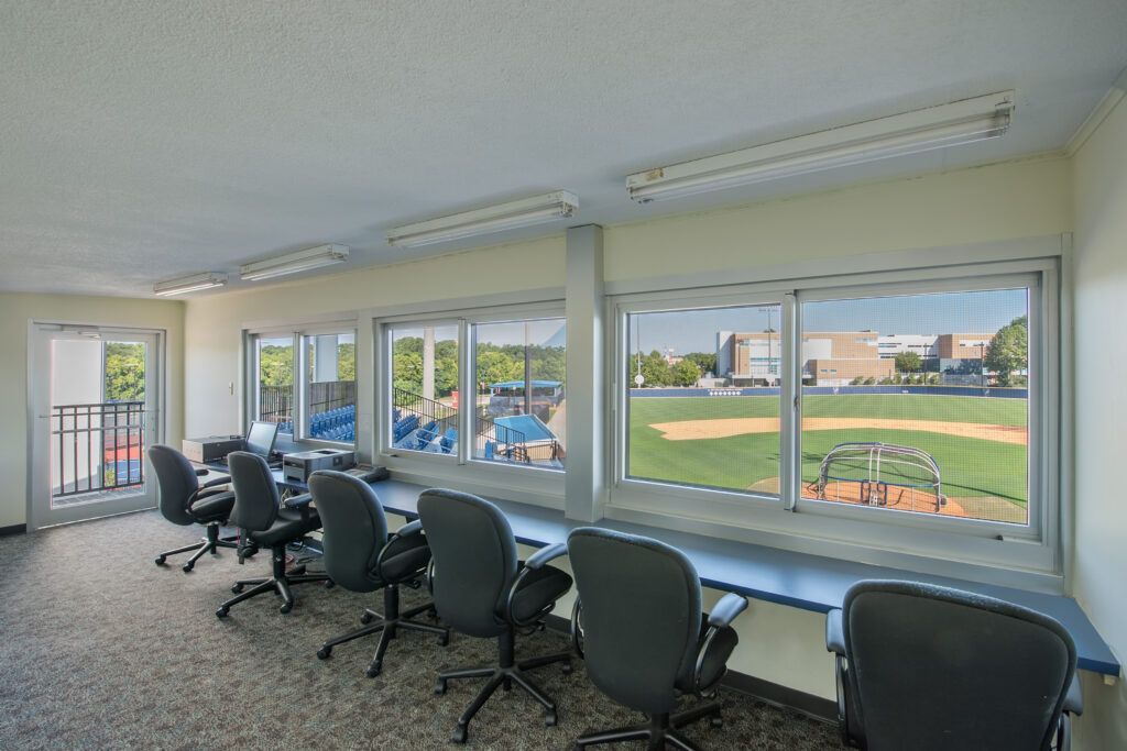 A room with a lot of windows and chairs and a view of a baseball field.