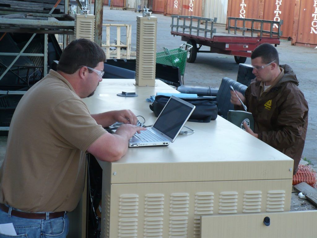 Two men are sitting at a table with a laptop and one has a ups logo on his jacket