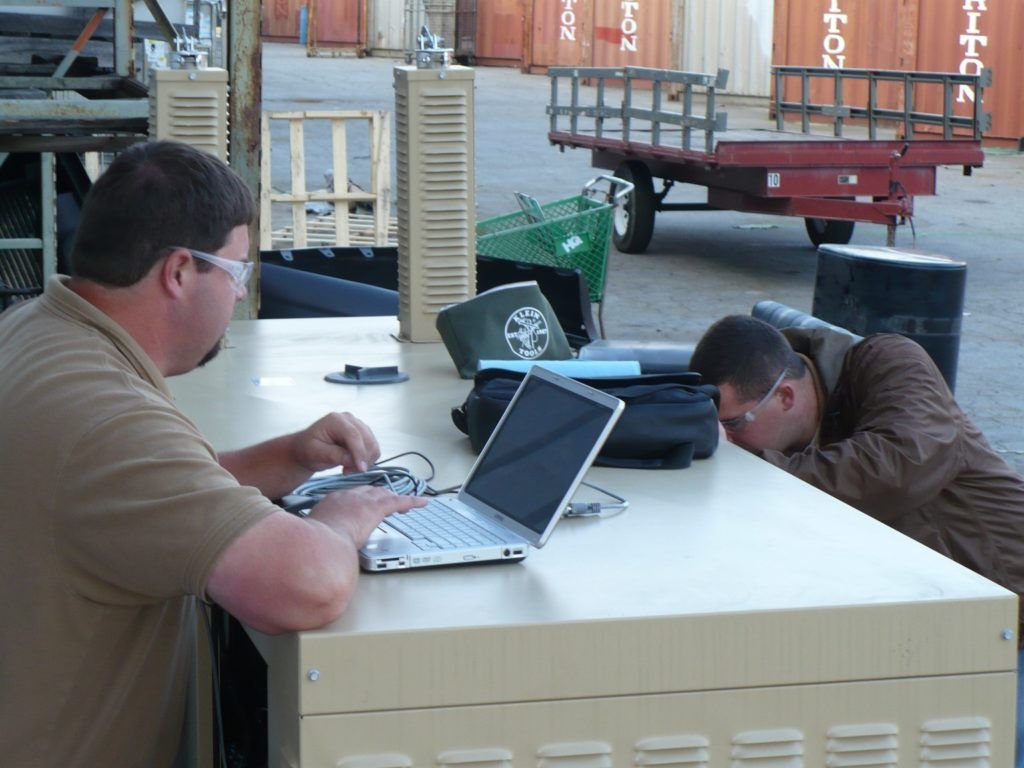 Two men are working on laptops in front of shipping containers that say cotton