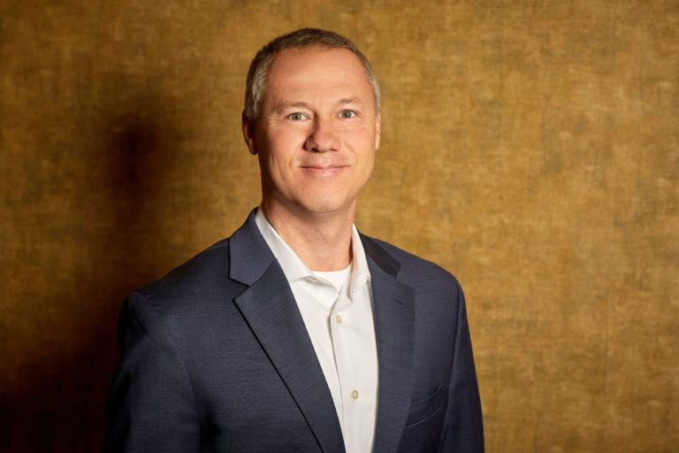 A man in a suit and white shirt is standing in front of a brown wall.