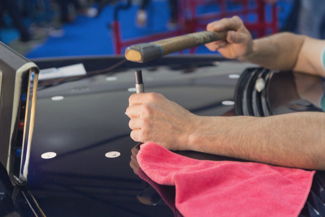 Person using a hammer to remove a dent from a car hood, working with a tool and cloth.