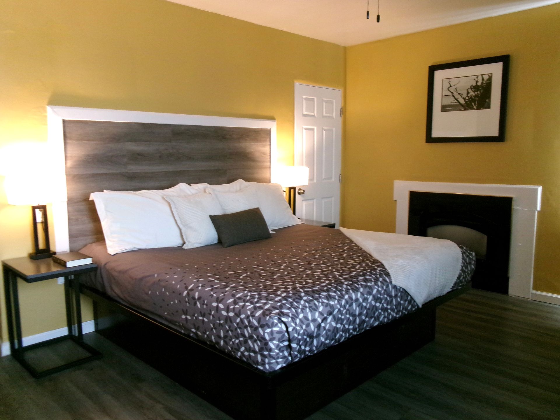 A bedroom with golden-yellow walls, a bed featuring a gray patterned quilt, and a dark fireplace on the right side.