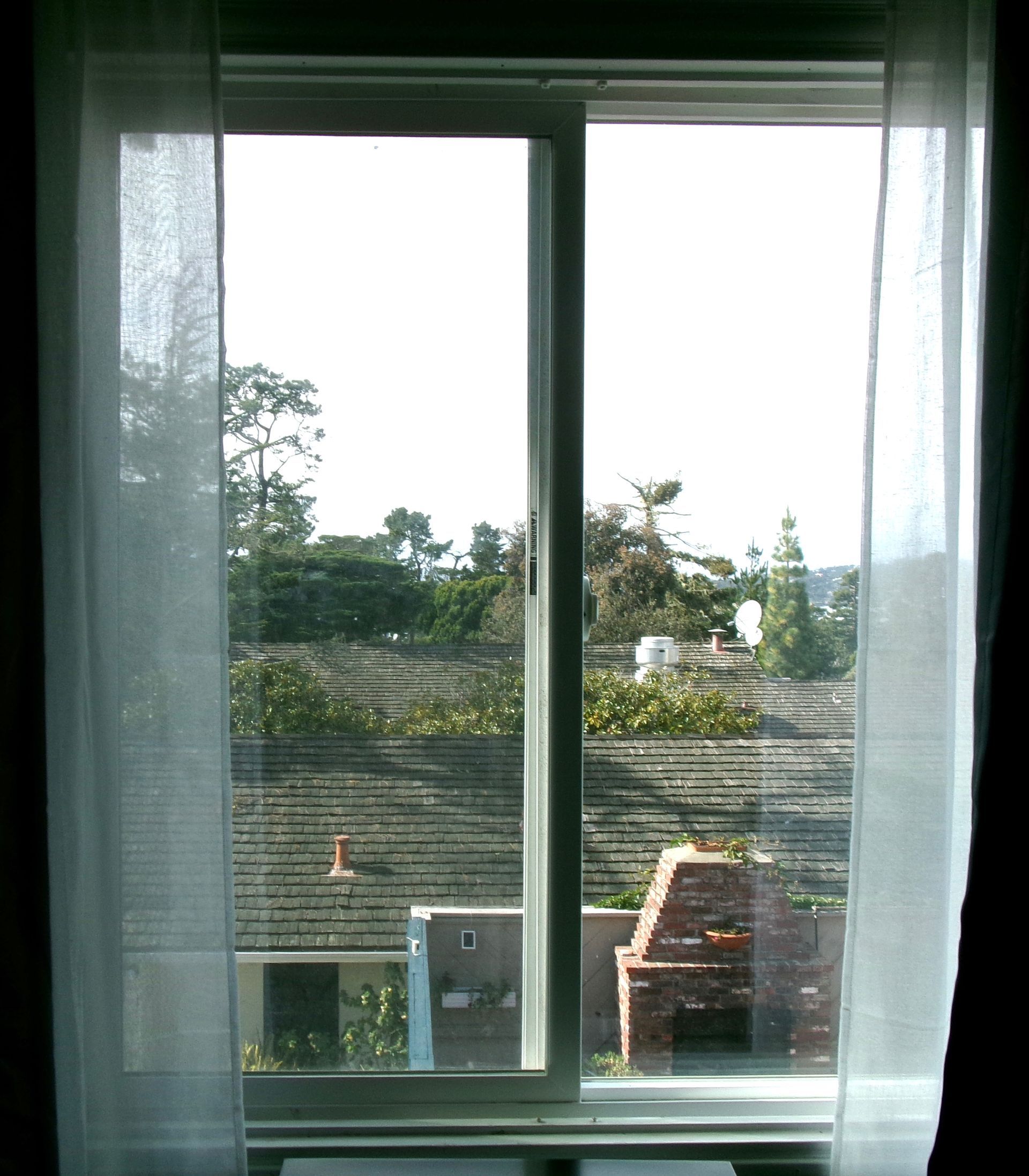 A view through a window with white sheer curtains, showing trees, rooftops, and a brick chimney in the daylight.
