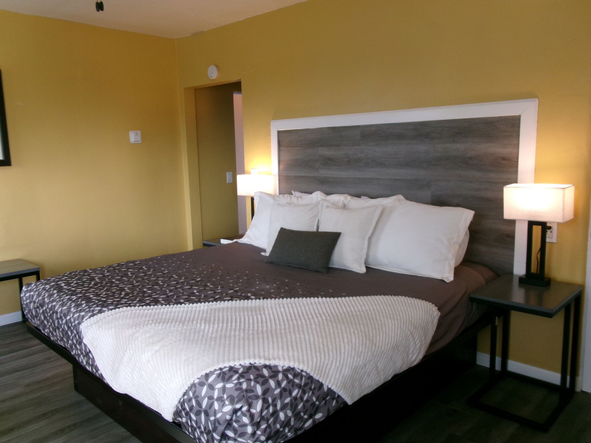 A modern bedroom with mustard-yellow walls, a bed with a gray wood-paneled headboard, white bedding, and two side tables.