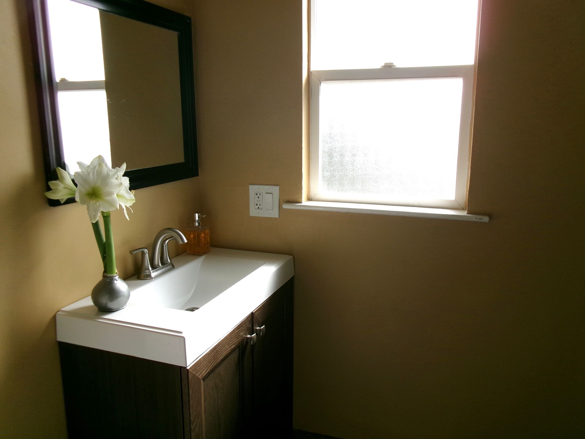 A vanity with a white sink and dark cabinet below a mirror and window in a room with tan walls and a vase of flowers.