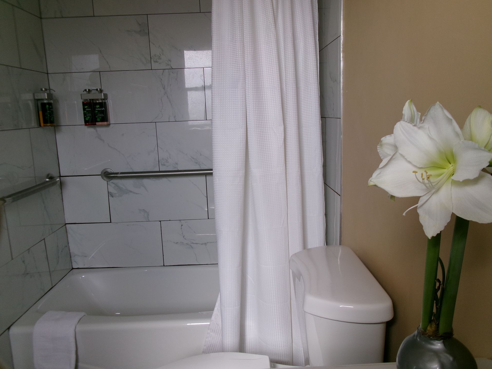 A bathroom with a white shower curtain, a tiled bathtub with a grab bar, and a vase of white amaryllis flowers.
