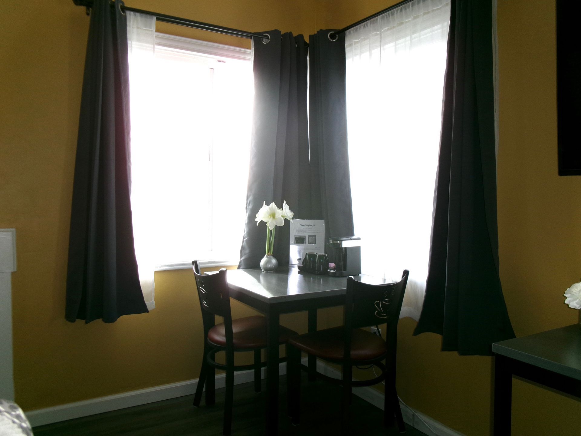 A small dining table with two chairs sits between two windows covered by dark curtains in a room with yellow walls.