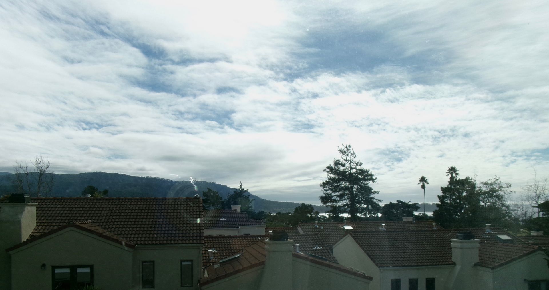 A view of neighborhood rooftops under a bright, cloudy sky with distant hills in the background.