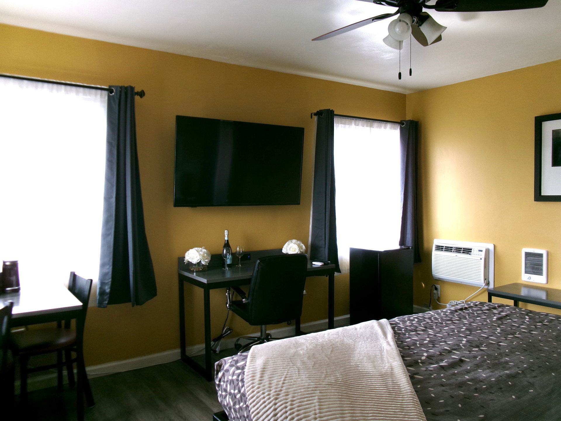 A bedroom with yellow walls, a dark-themed bed, a desk with a chair under a wall-mounted TV, and two windows with curtains.