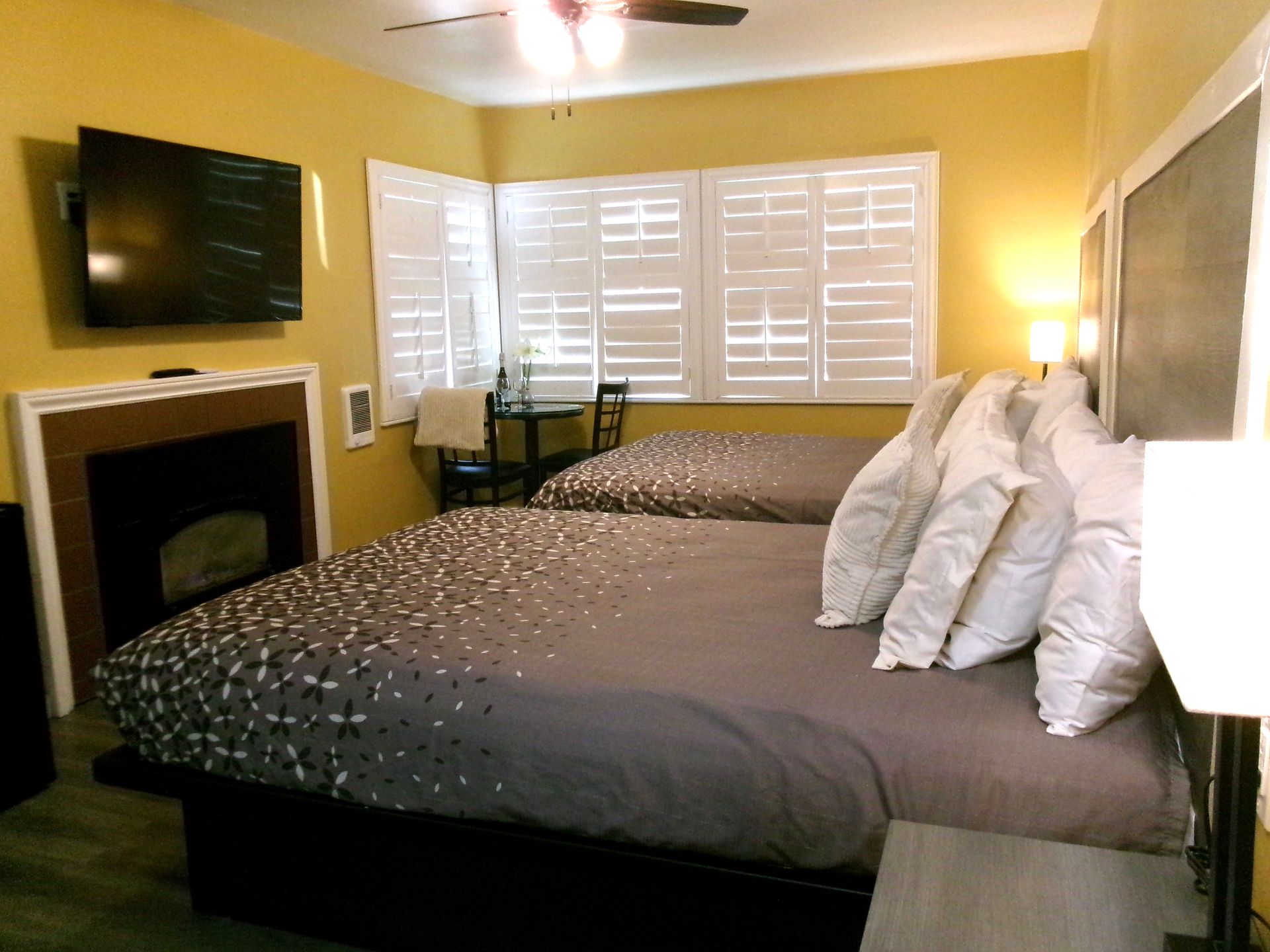 A hotel room with two beds, a fireplace, a mounted television, and a window with white shutters.