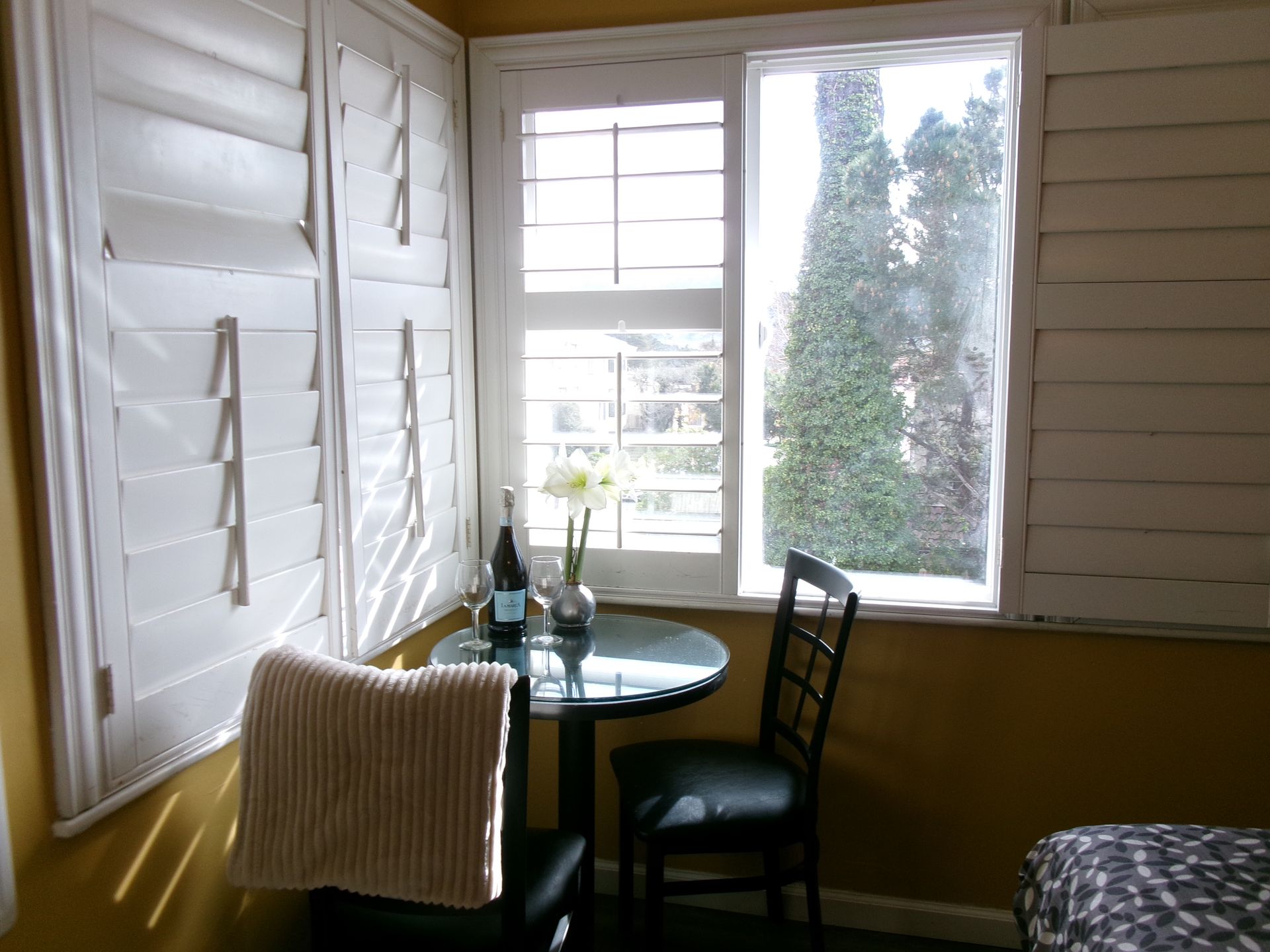 A small glass bistro table and two chairs sit by a window with white plantation shutters and yellow walls.