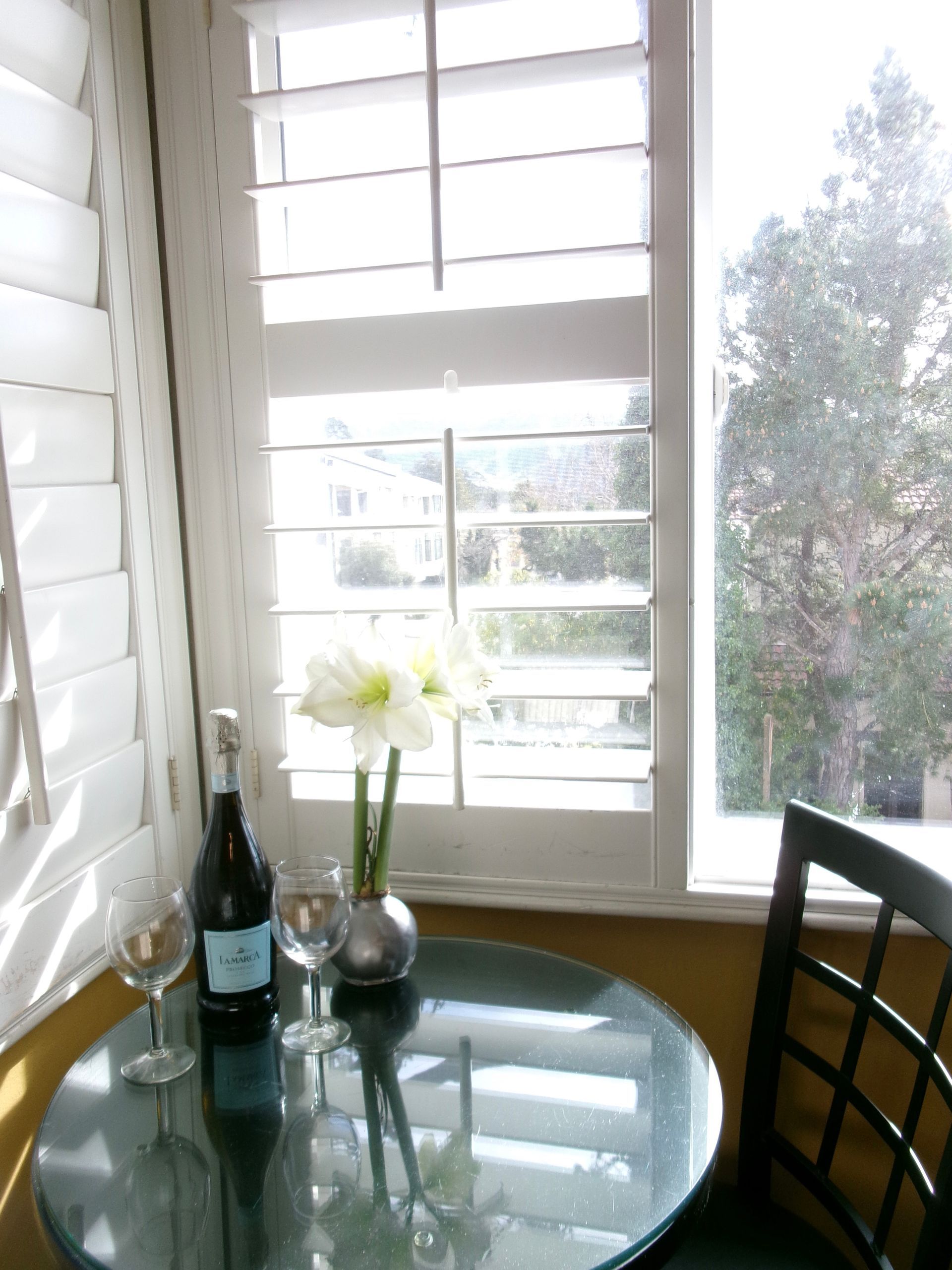 A wine bottle and two glasses next to a vase with white flowers on a glass-topped table in front of a shuttered window.