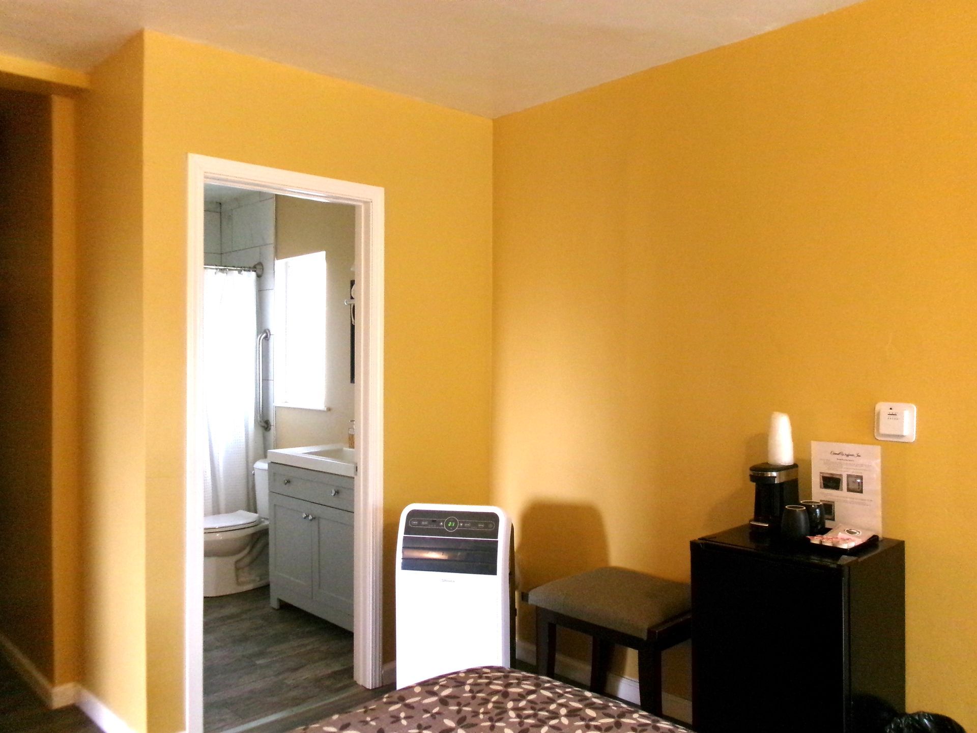 A yellow-walled room with a doorway showing a bathroom, a portable air conditioner, and a mini-fridge with coffee items.