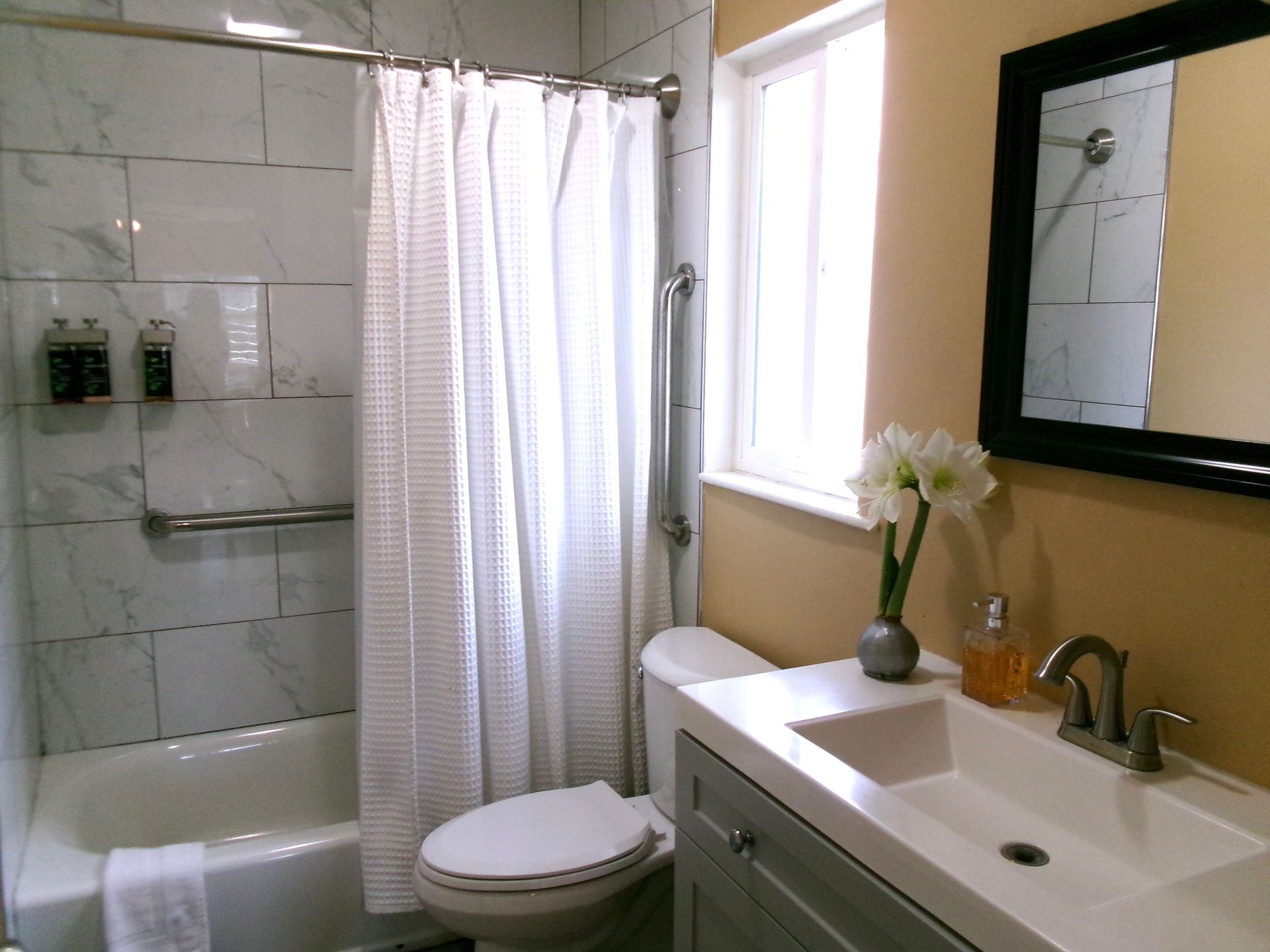 A modern bathroom featuring a white marble-tiled tub/shower, a grey vanity with a white sink, and a dark-framed mirror.