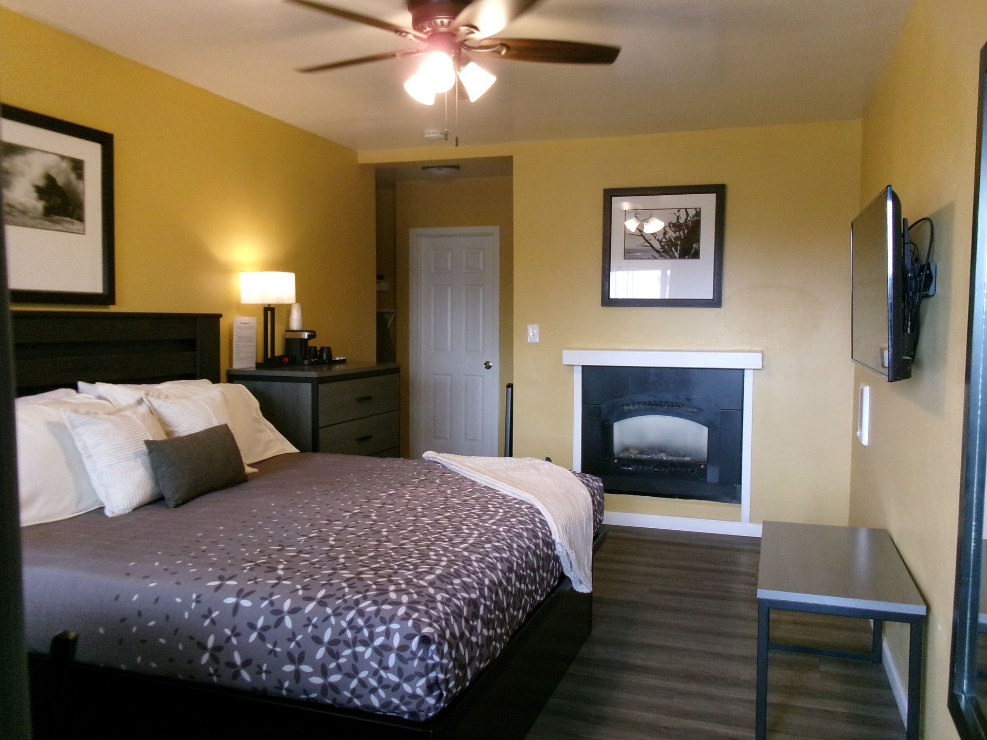 A hotel bedroom featuring a bed with patterned bedding, a fireplace, a television on the wall, and yellow walls.