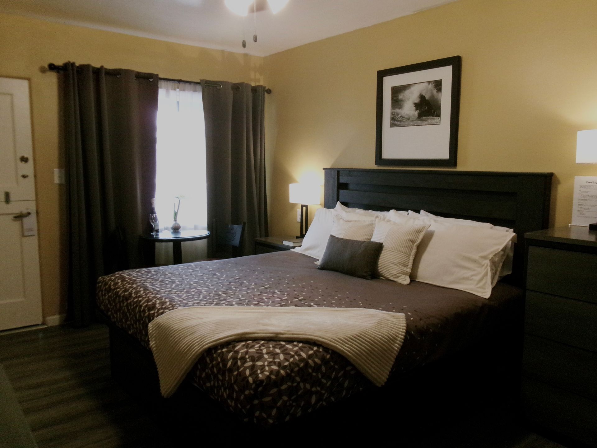 A bedroom featuring a bed with patterned linens, a dark headboard, two lamps, a framed picture, and floor-length curtains.