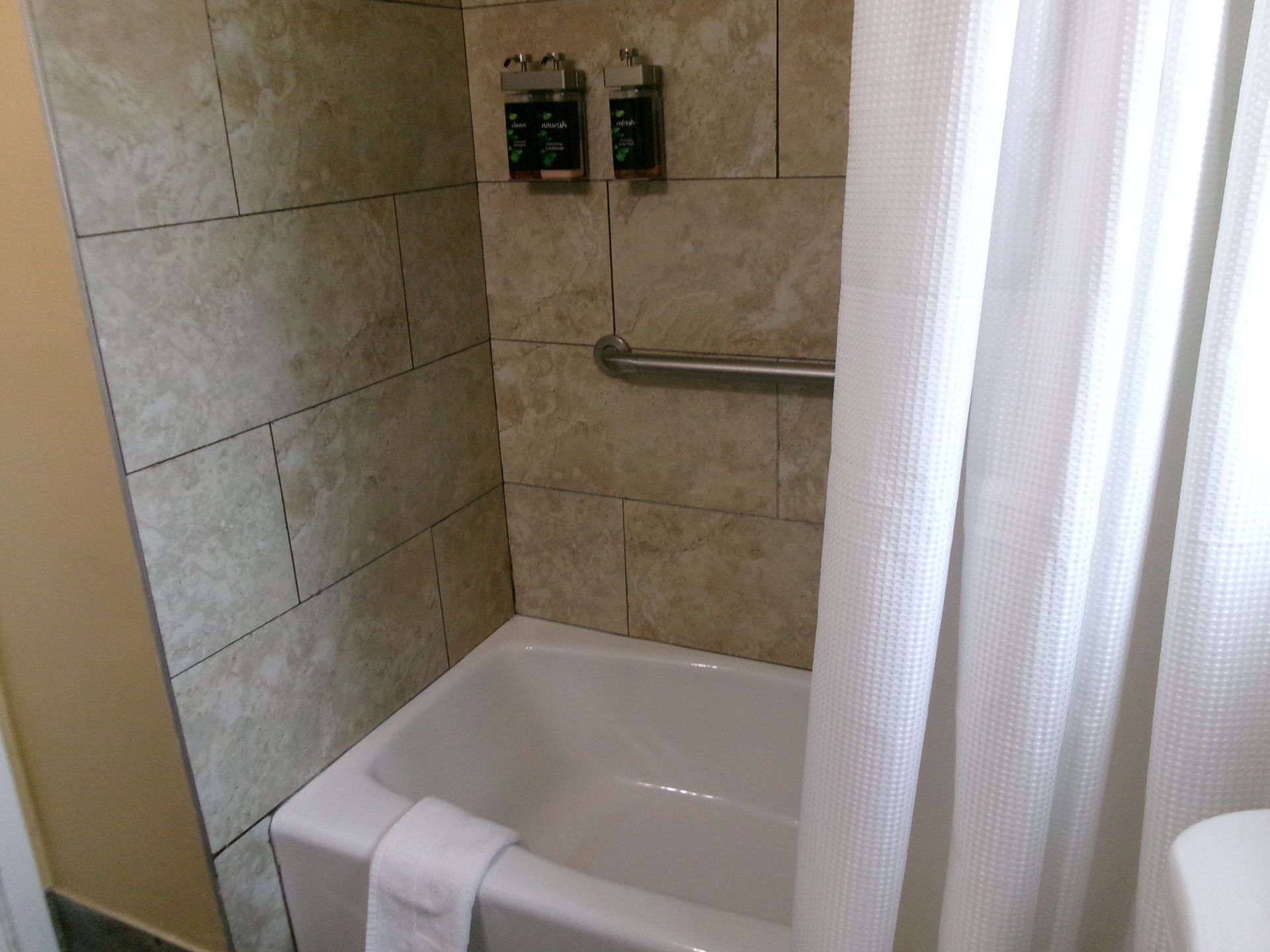 A hotel bathroom shower-tub combination with light-colored tile walls, a silver grab bar, and wall-mounted dispensers.