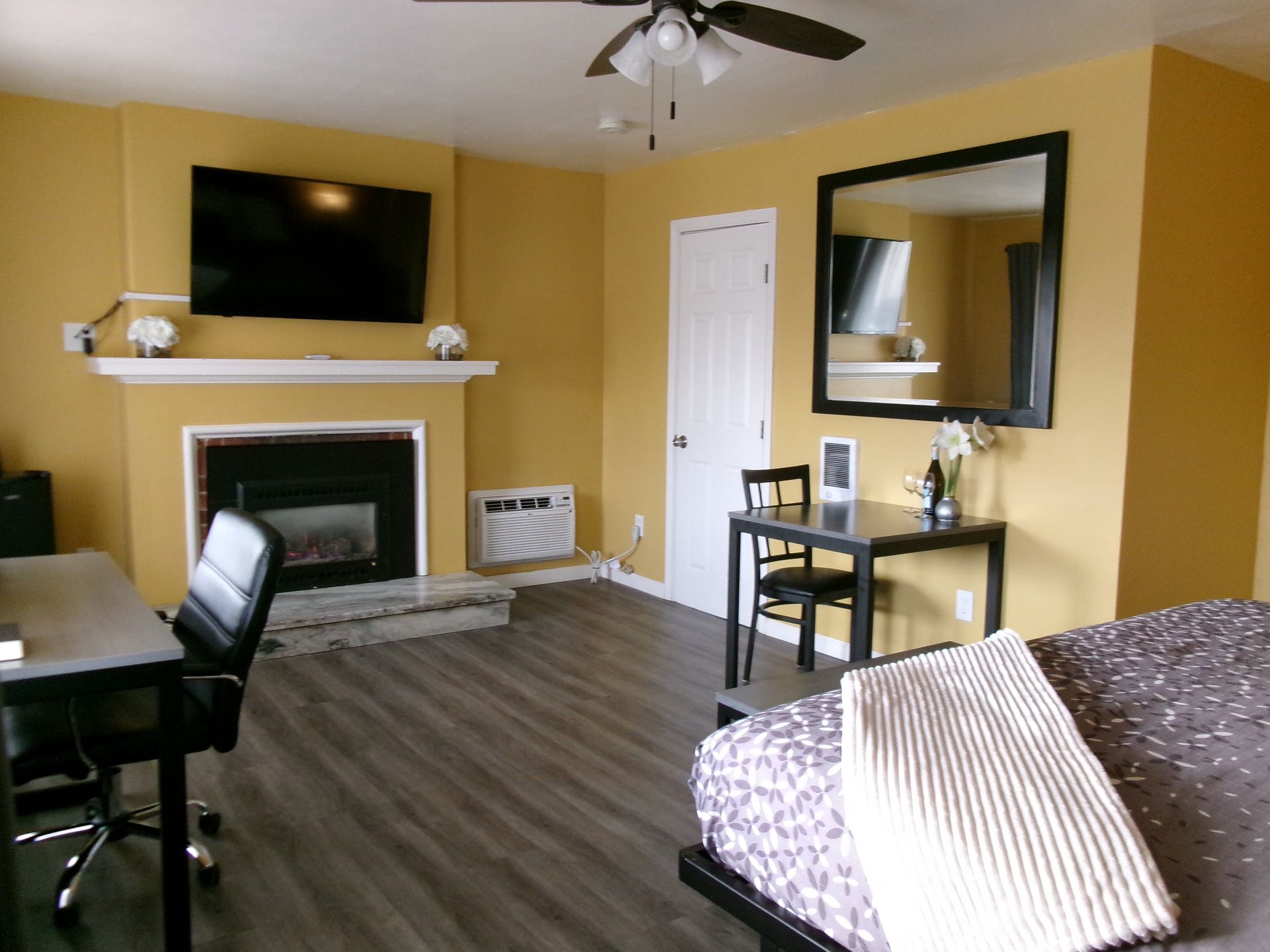 A hotel room with yellow walls, a fireplace with a mounted TV, a small dining table, and a bed with a patterned cover.
