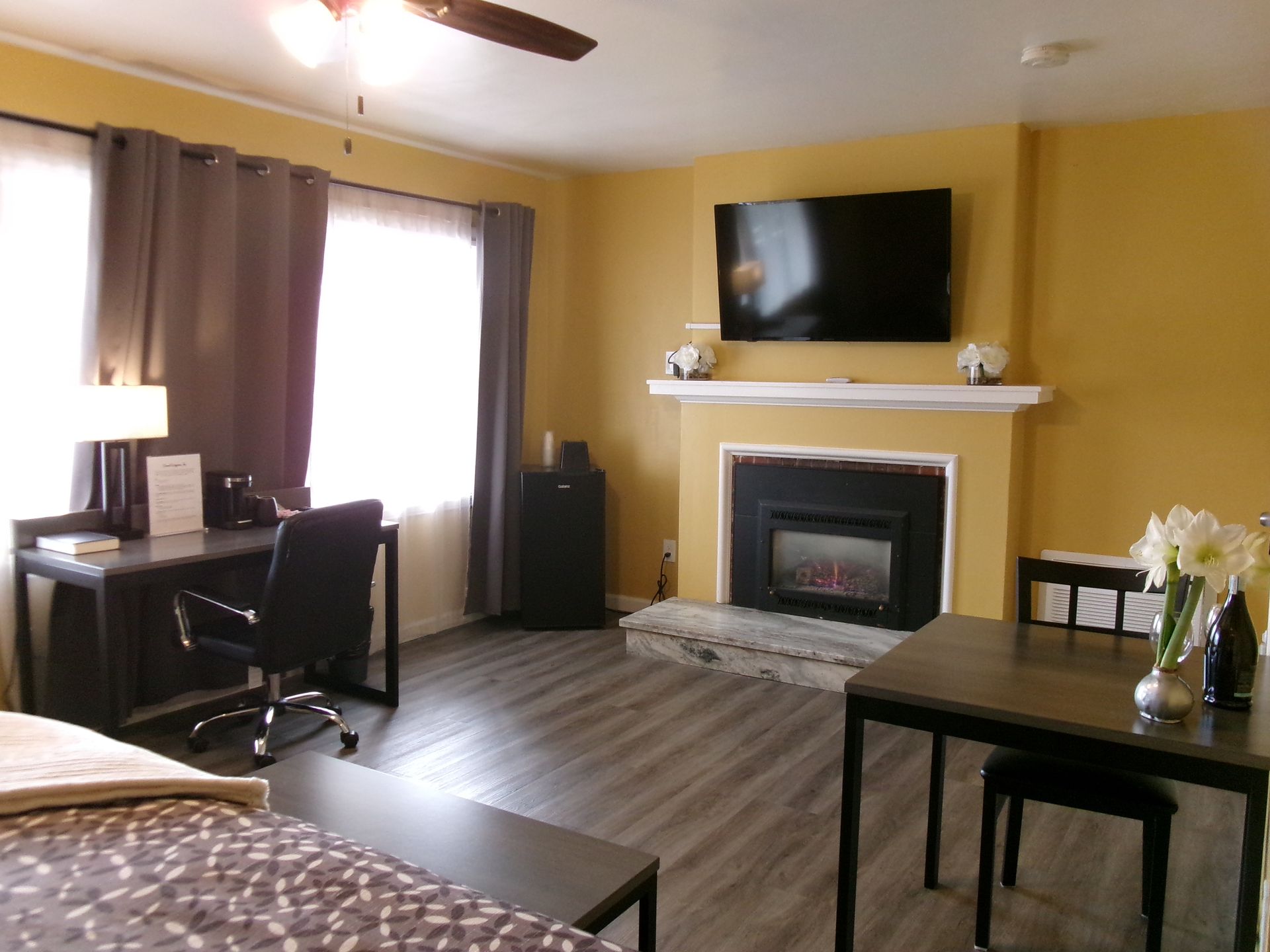 A hotel room with yellow walls, gray floors, a desk, a dining table, and a fireplace with a mounted television.
