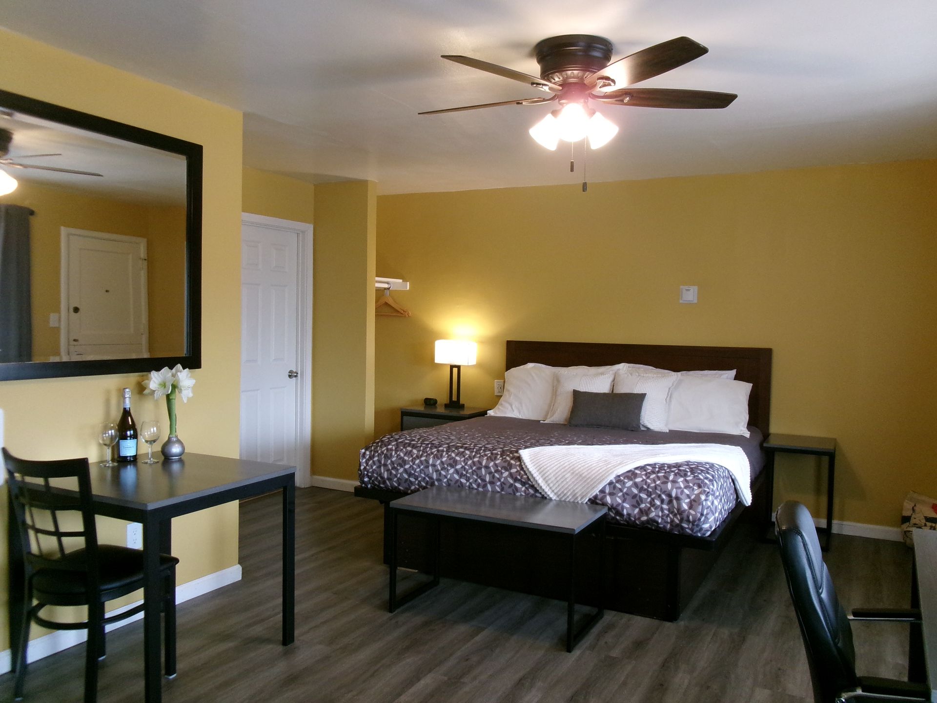A hotel room with a yellow-walled bedroom featuring a king bed, a ceiling fan, and a dining table with a chair.