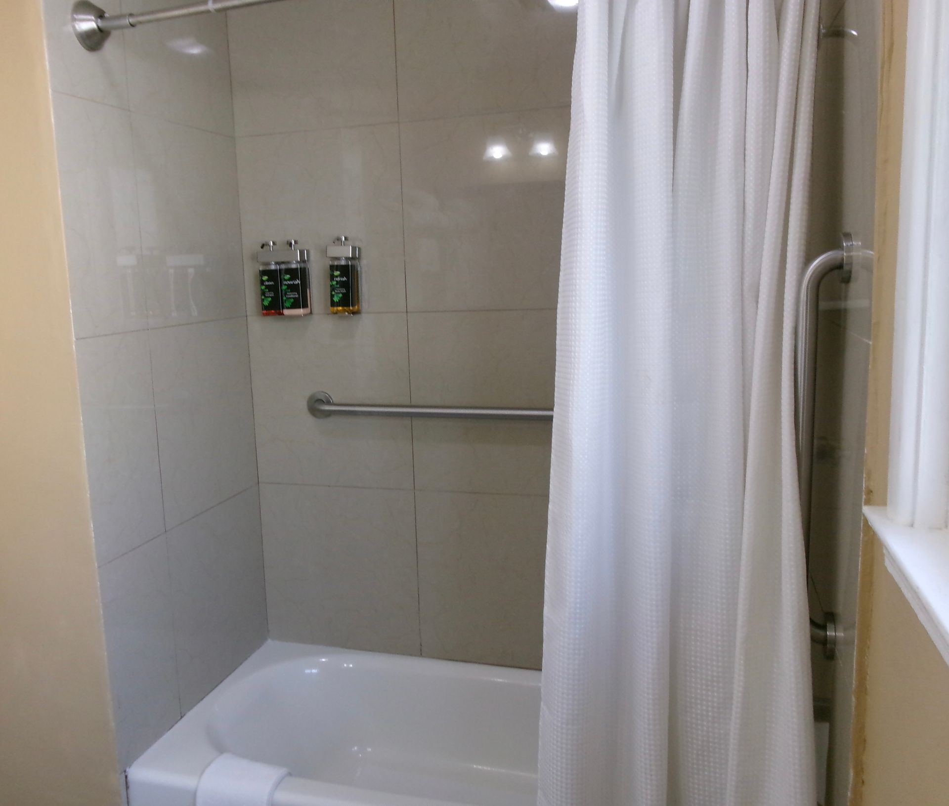 A white bathtub and shower with beige tiled walls, a curtain, two grab bars, and mounted soap dispensers.