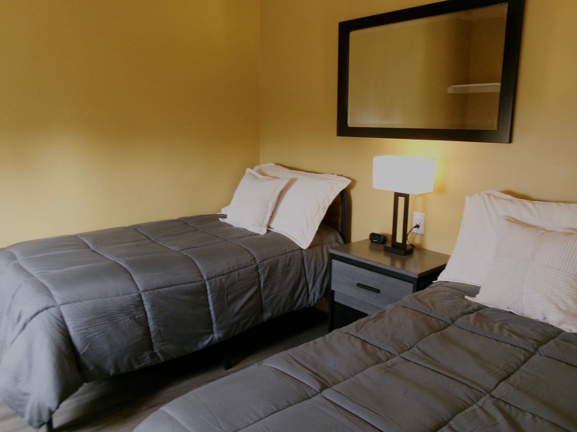 Two twin beds with gray comforters separated by a nightstand with a lamp, with a black-framed mirror hanging above.