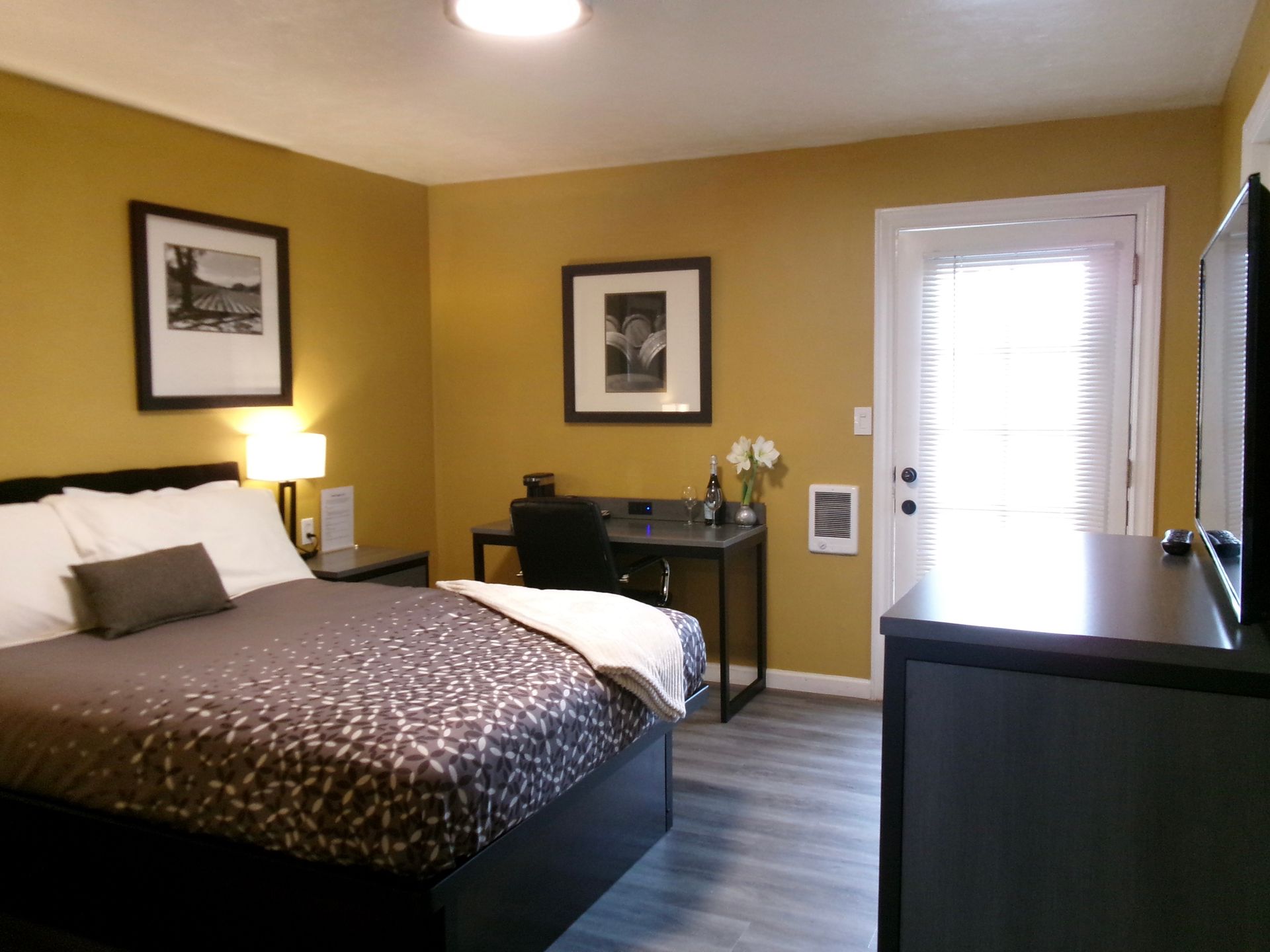 A hotel room featuring a bed with patterned bedding, a desk with a chair, and a door leading outside.