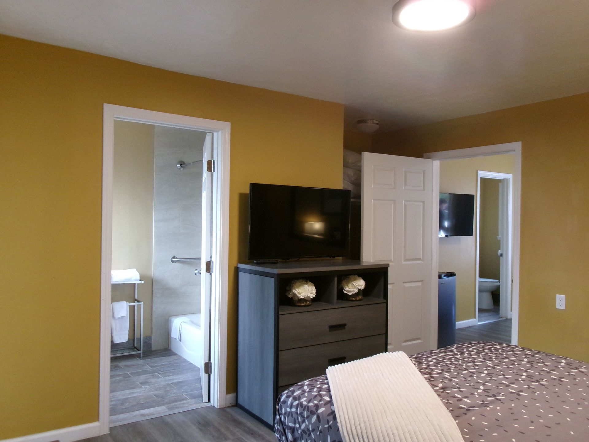 A hotel room with yellow walls, a dresser holding a TV, a white-blanketed bed, and two open doorways to bathrooms.