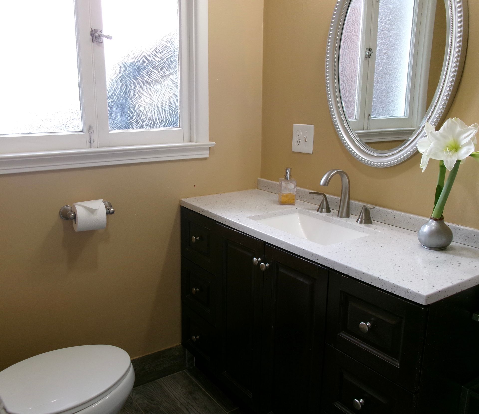 A bathroom featuring a dark wood vanity with a white speckled countertop, oval silver mirror, and a white toilet.
