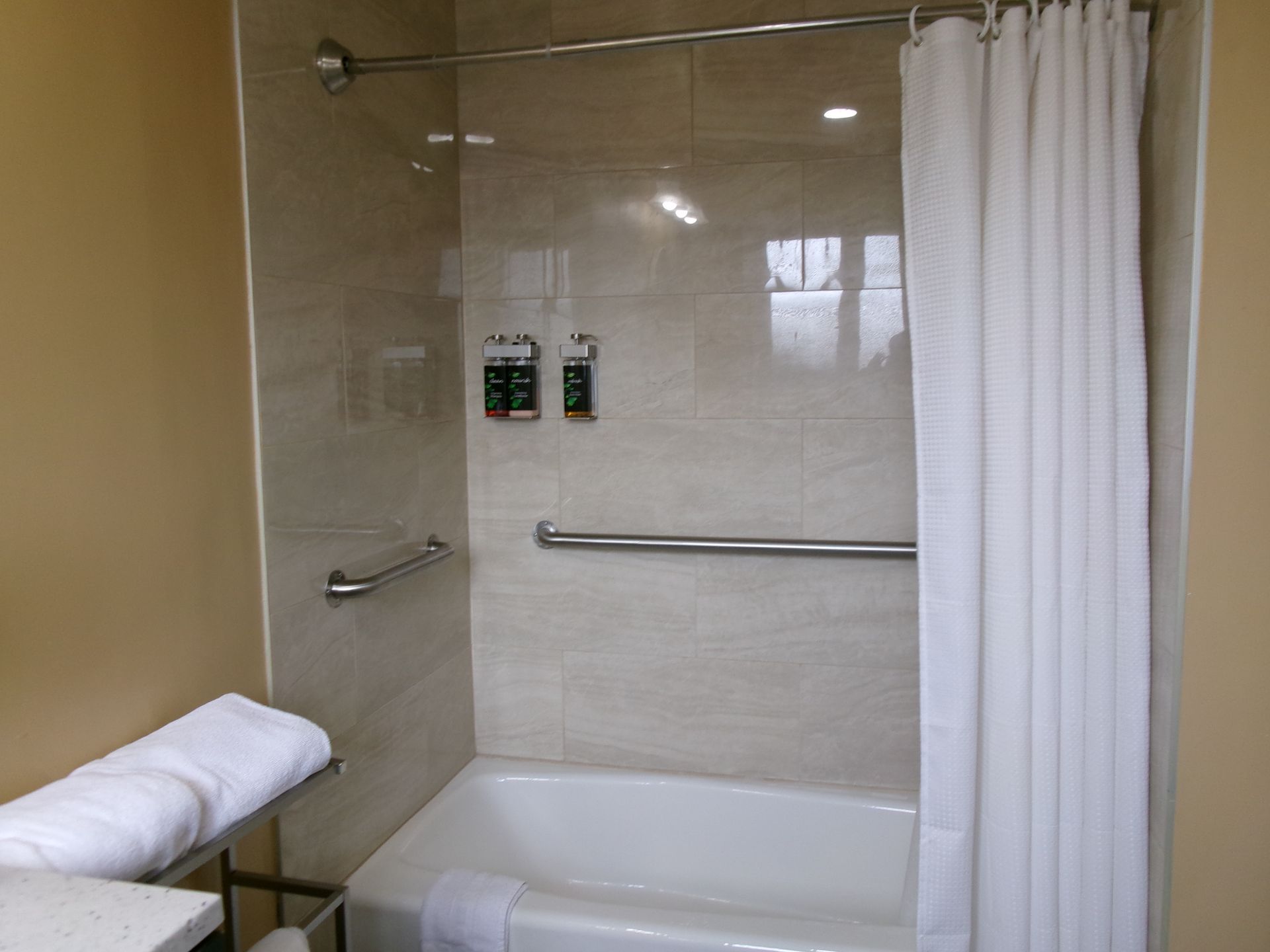 A bathroom bathtub and shower combination with tan tile walls, grab bars, soap dispensers, and a white shower curtain.