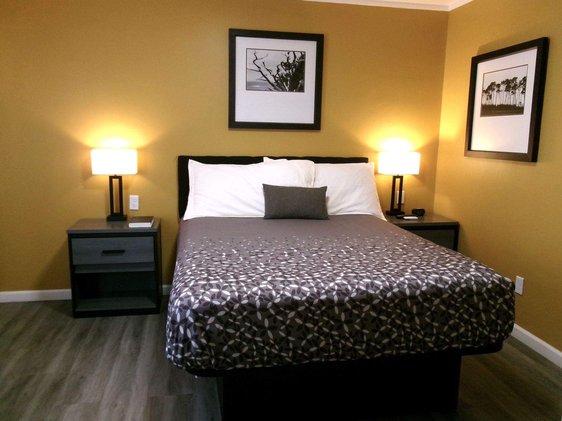 A bedroom with a bed featuring a patterned gray quilt, two nightstands with lamps, and two framed prints on gold walls.