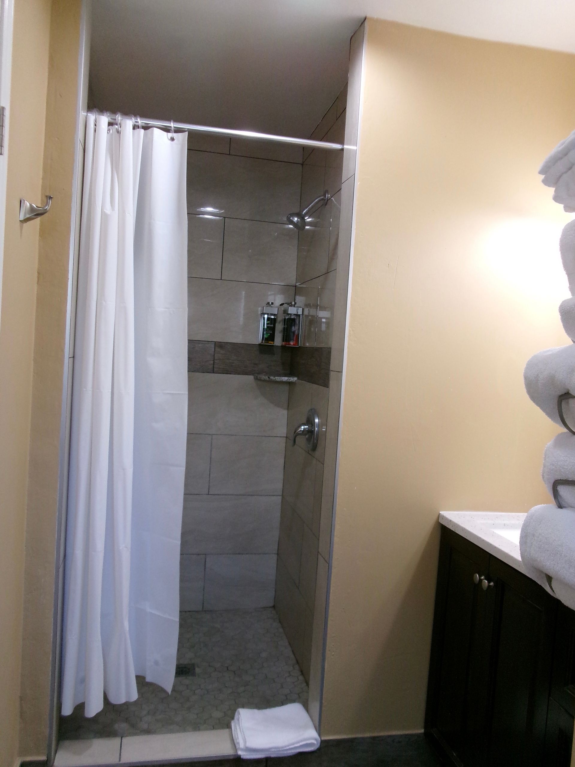 A tiled shower stall with a white curtain, adjacent to a brown vanity with a stack of white towels.