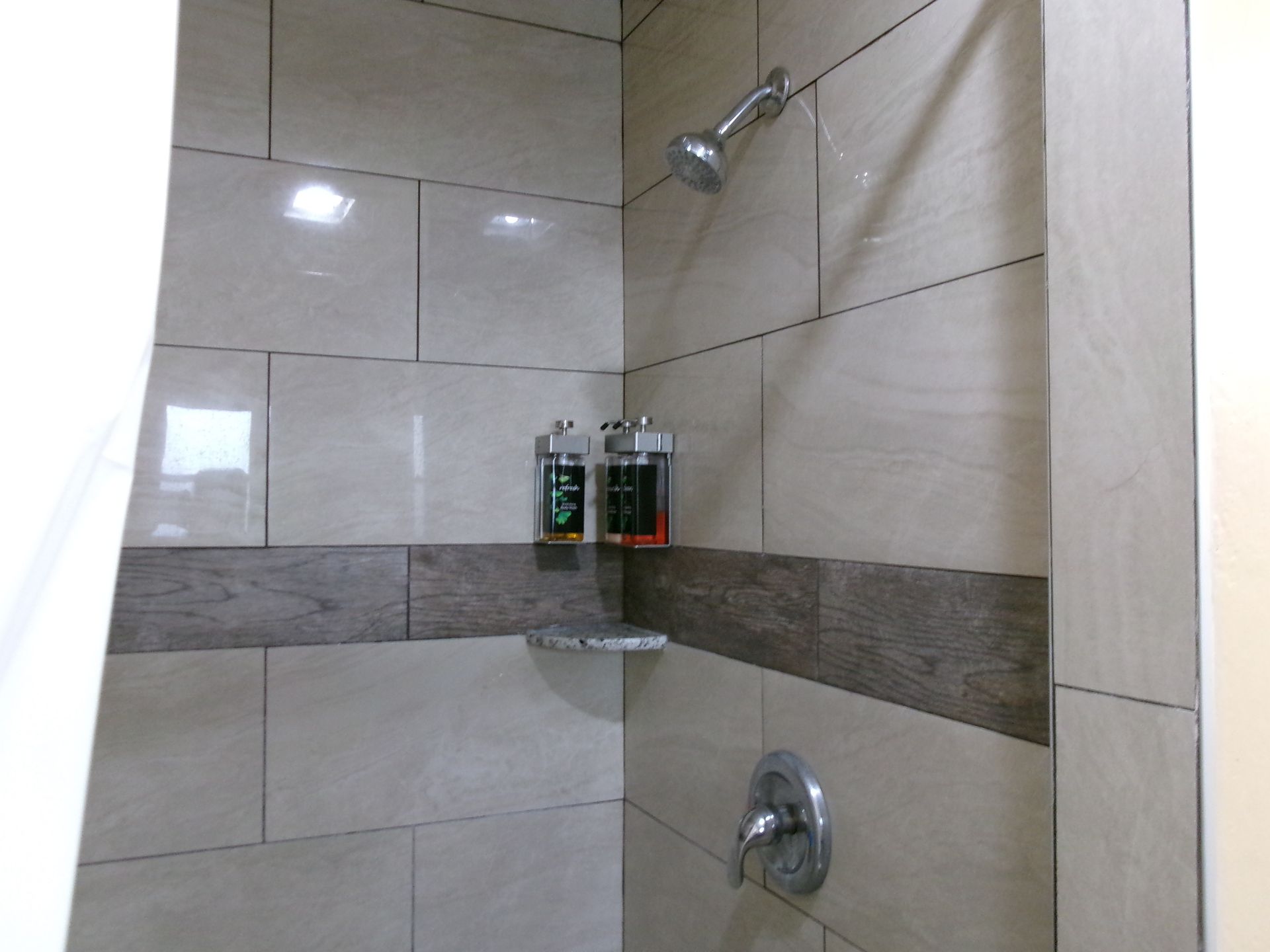 A shower stall with tan tiled walls, a horizontal dark wood-look accent band, a shower head, and two wall-mounted dispensers.