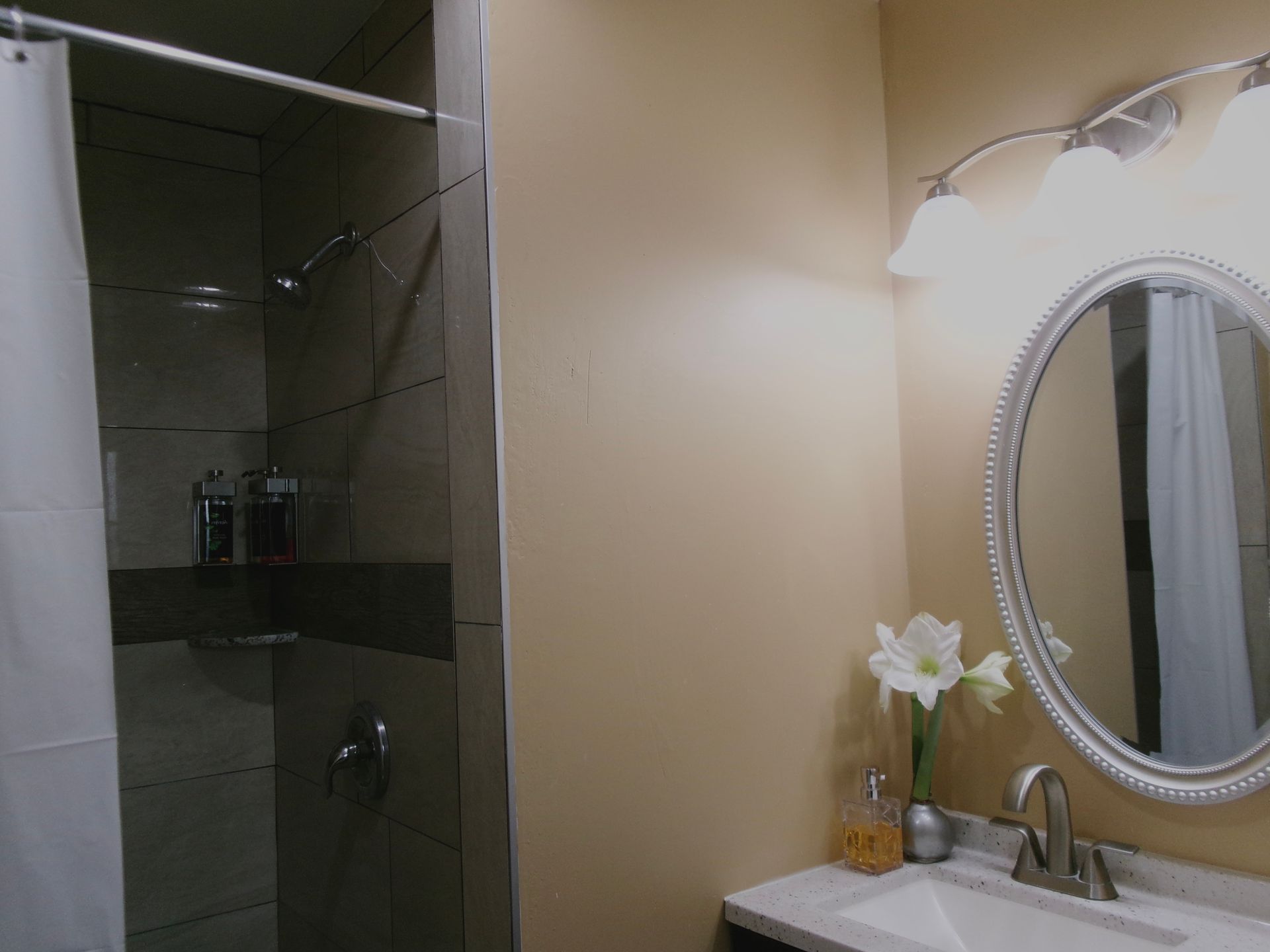 A bathroom featuring a tiled shower with a white curtain, an oval mirror, light fixtures, and a sink with a floral vase.