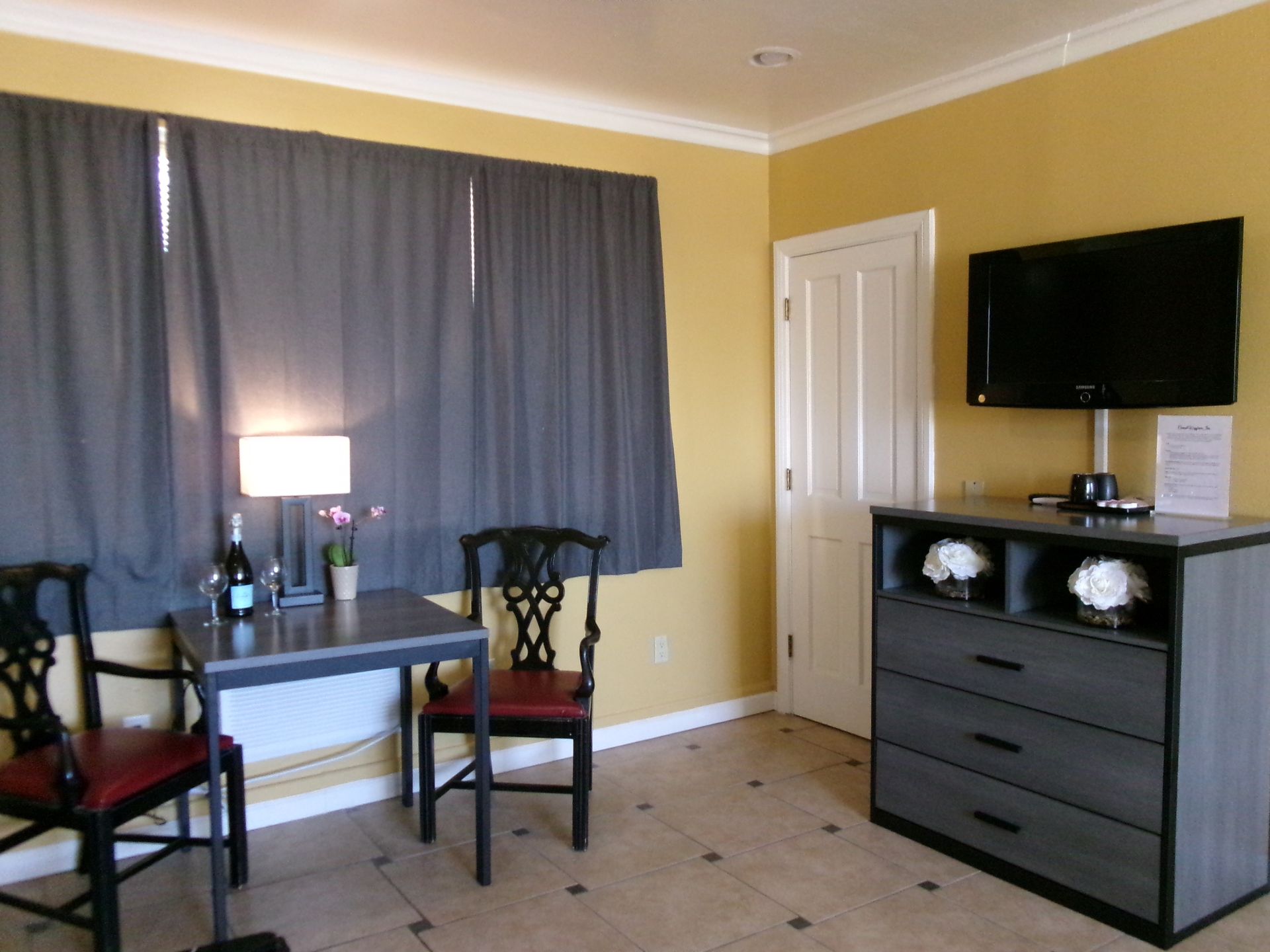 A room with yellow walls, a dining table with two chairs, a lamp, and a dresser with a wall-mounted television.
