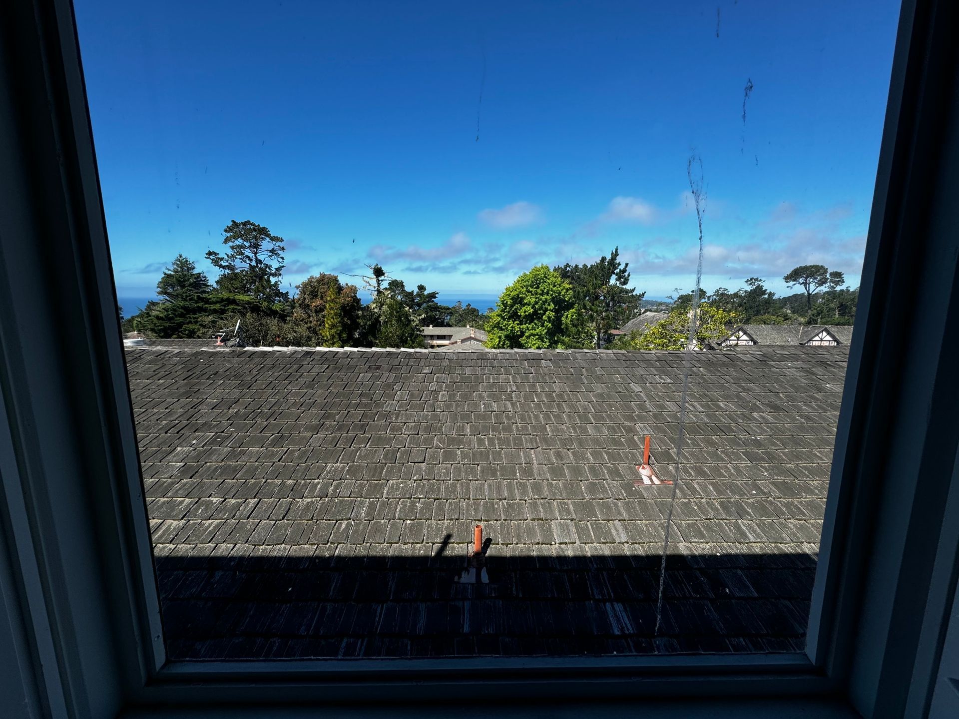 A view through a window showing a gray shingled roof under a bright blue sky with trees in the distance.