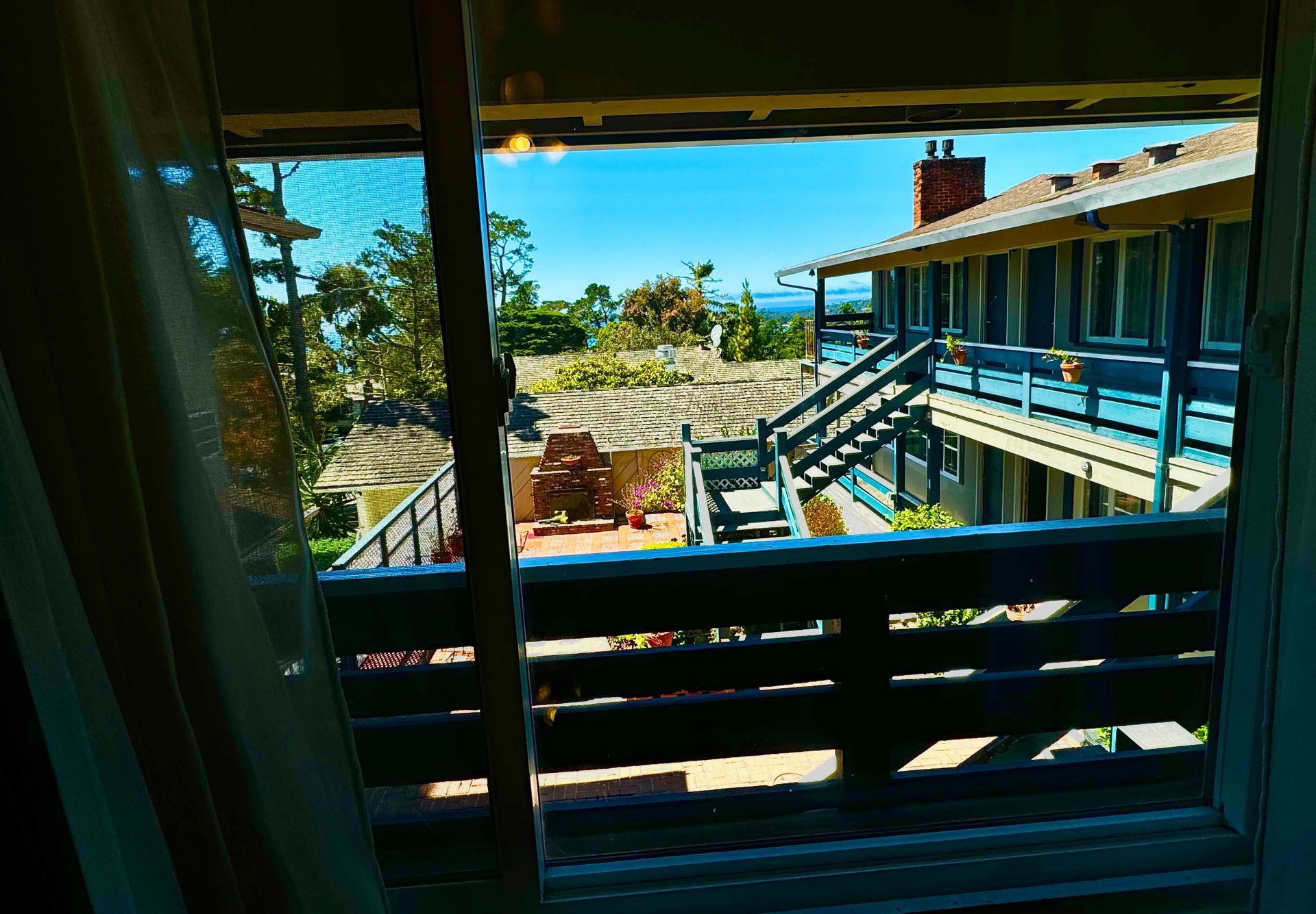 View from a window overlooking a sunny motel balcony with stairs, a patio area, and distant greenery under a clear sky.
