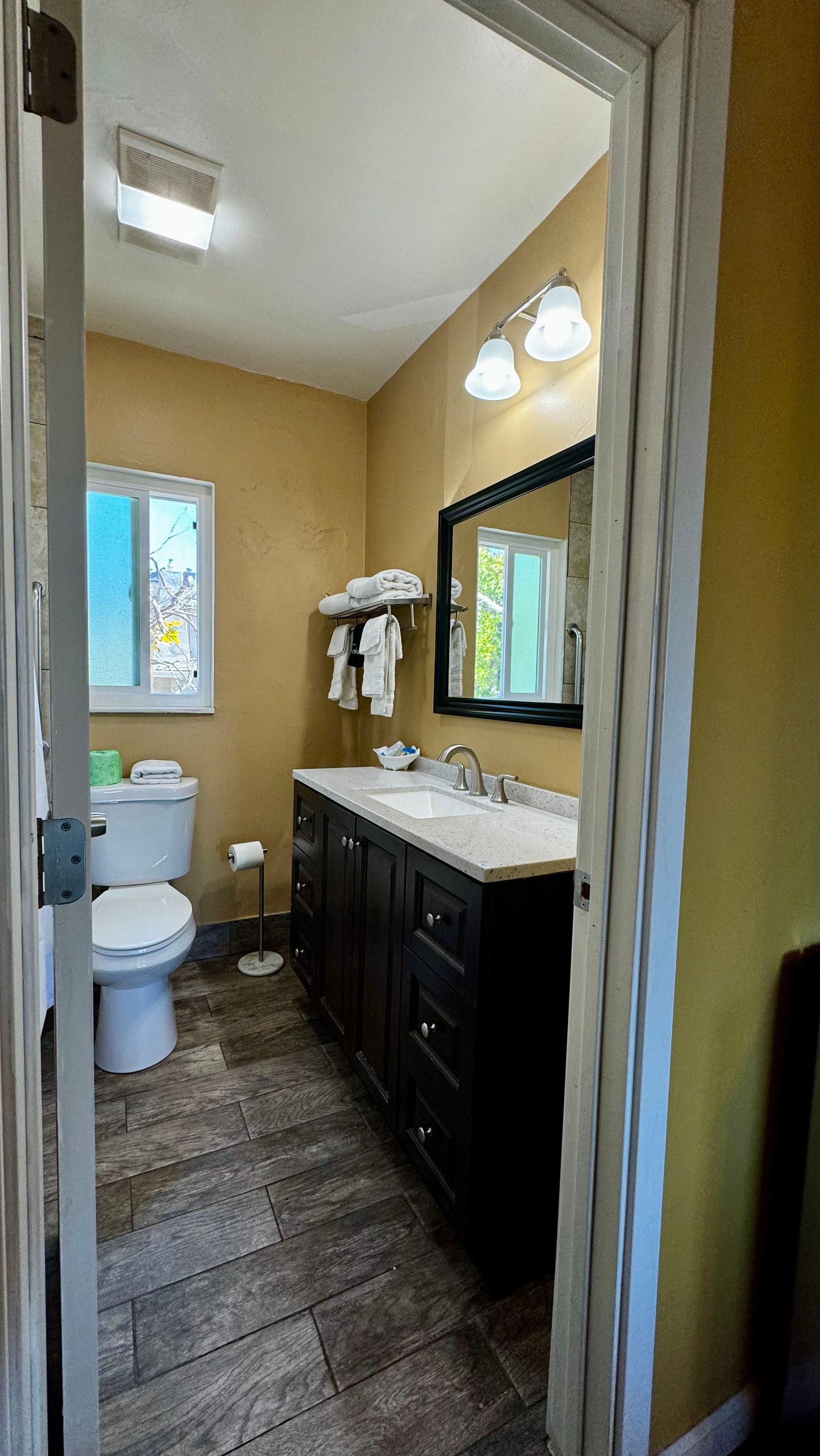 A small bathroom with tan walls, a dark wood vanity, a mirror, and a white toilet on a textured brown tiled floor.